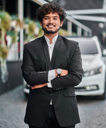 A member of the Intellgus Team, dressed in a black suit and white shirt, stands confidently with arms crossed in front of a white car, complemented by greenery and a building in the background.