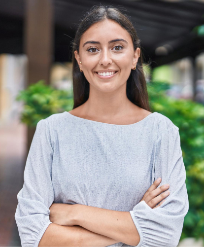 A person with long dark hair, symbolizing the confident spirit of the Intellgus Team, stands outdoors with arms crossed, smiling. They are wearing a light gray top against a backdrop of blurred greenery and a walkway.