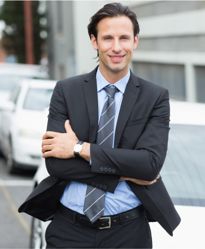 A smiling man in a suit and tie stands outdoors with arms crossed, leaning against a white car. He has dark hair and is wearing a wristwatch. The background includes blurred buildings and a row of cars.