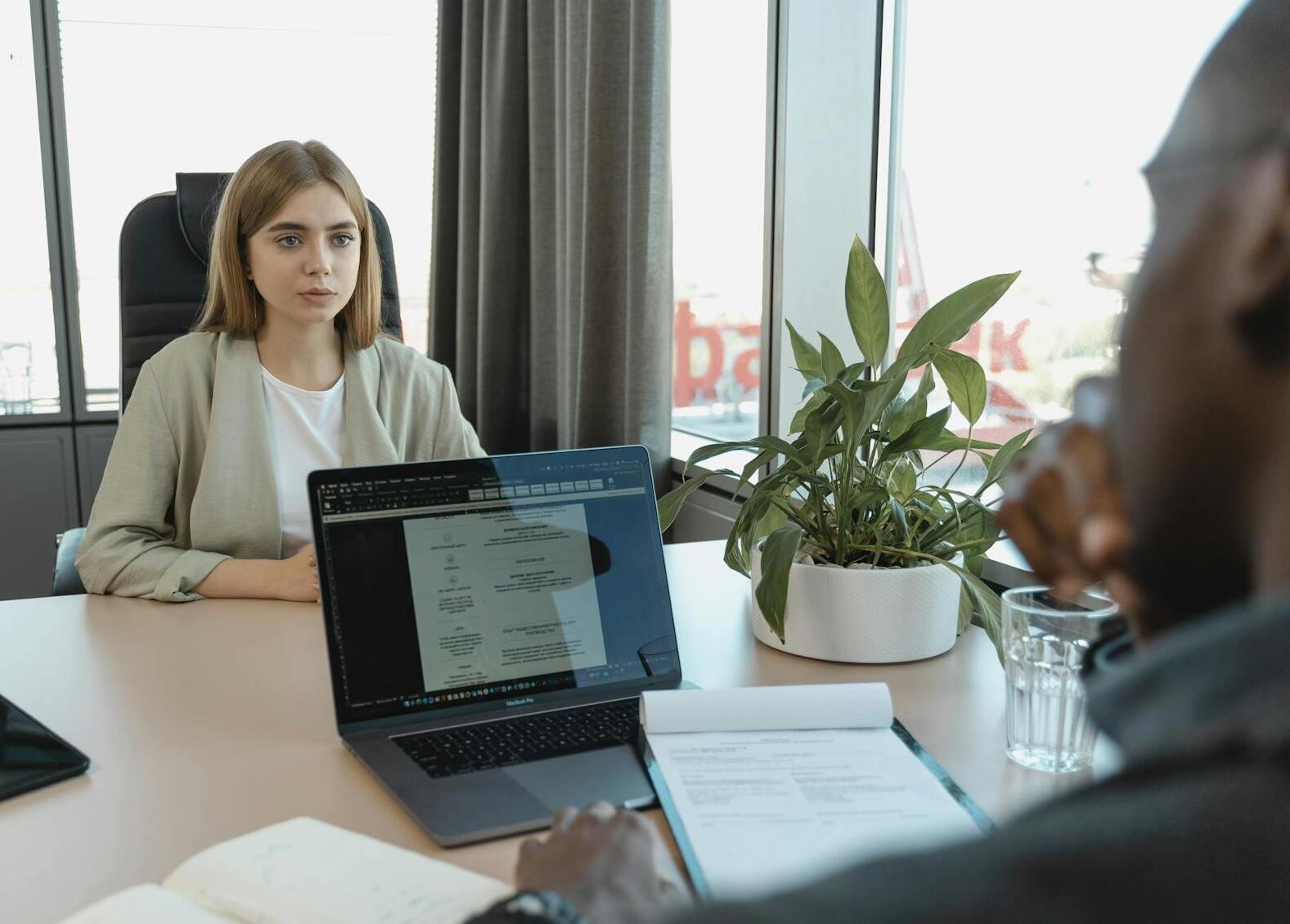 A woman sits across a desk from a man in a modern office, discussing best outsourced accounting services. A laptop with an open document, alongside a notebook and potted plant, lie on the desk. Both appear deeply engaged in their conversation about optimizing financial strategies.