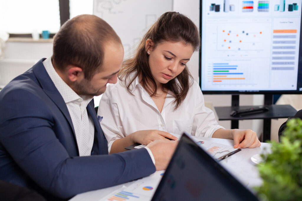 A man and a woman are sitting at a desk reviewing documents with graphs, likely analyzing data for tax audit services. A laptop is open in the foreground, and a screen displaying charts is visible in the background. They appear to be discussing work in an office setting.