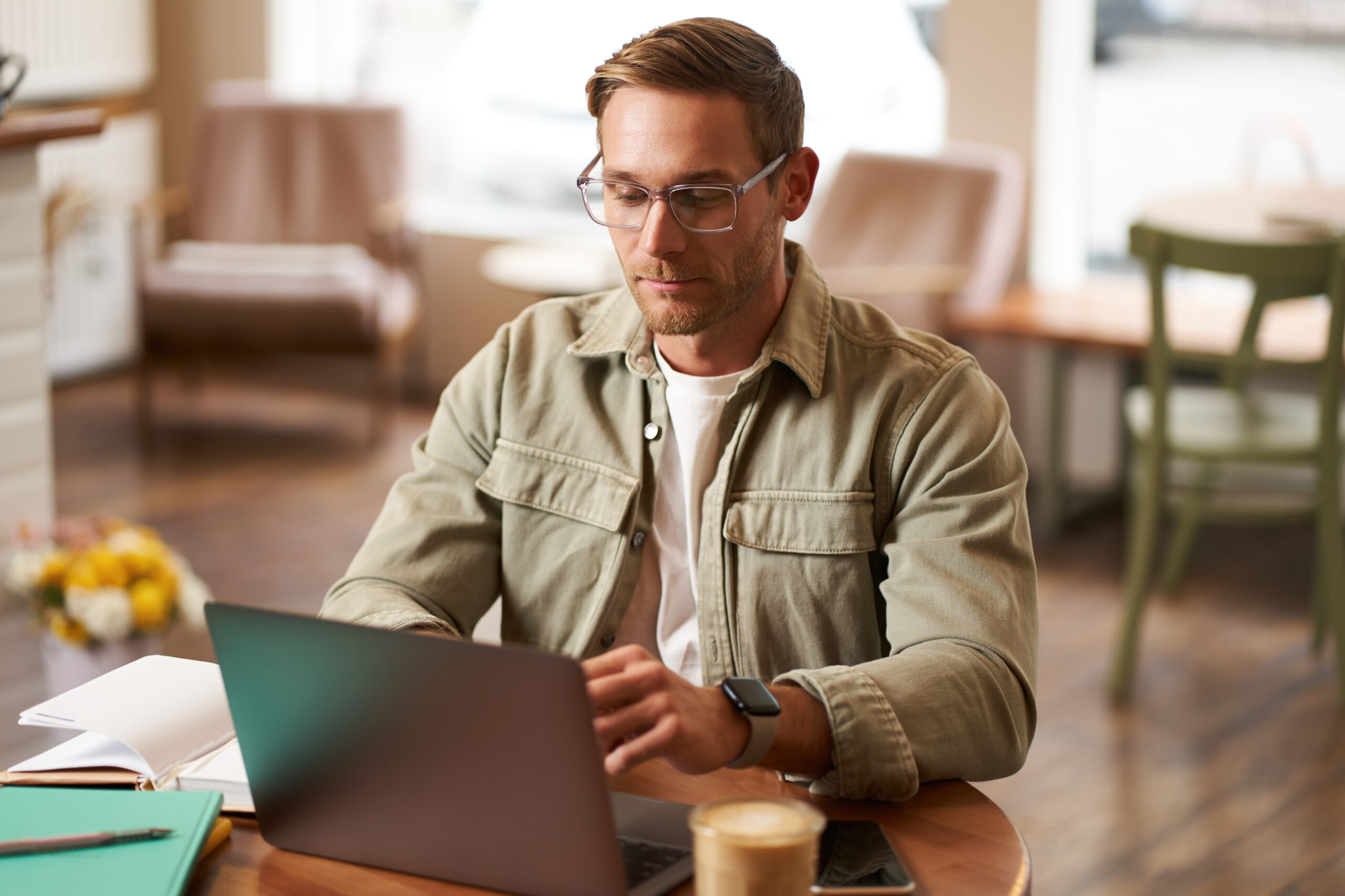 A man with glasses sits at a wooden table in a cozy cafe, focusing on outsourced bookkeeping tasks on his laptop. He wears a light green jacket and a smartwatch. The table holds a notebook, pen, and latte. Blurred chairs and flowers are softly visible in the background.