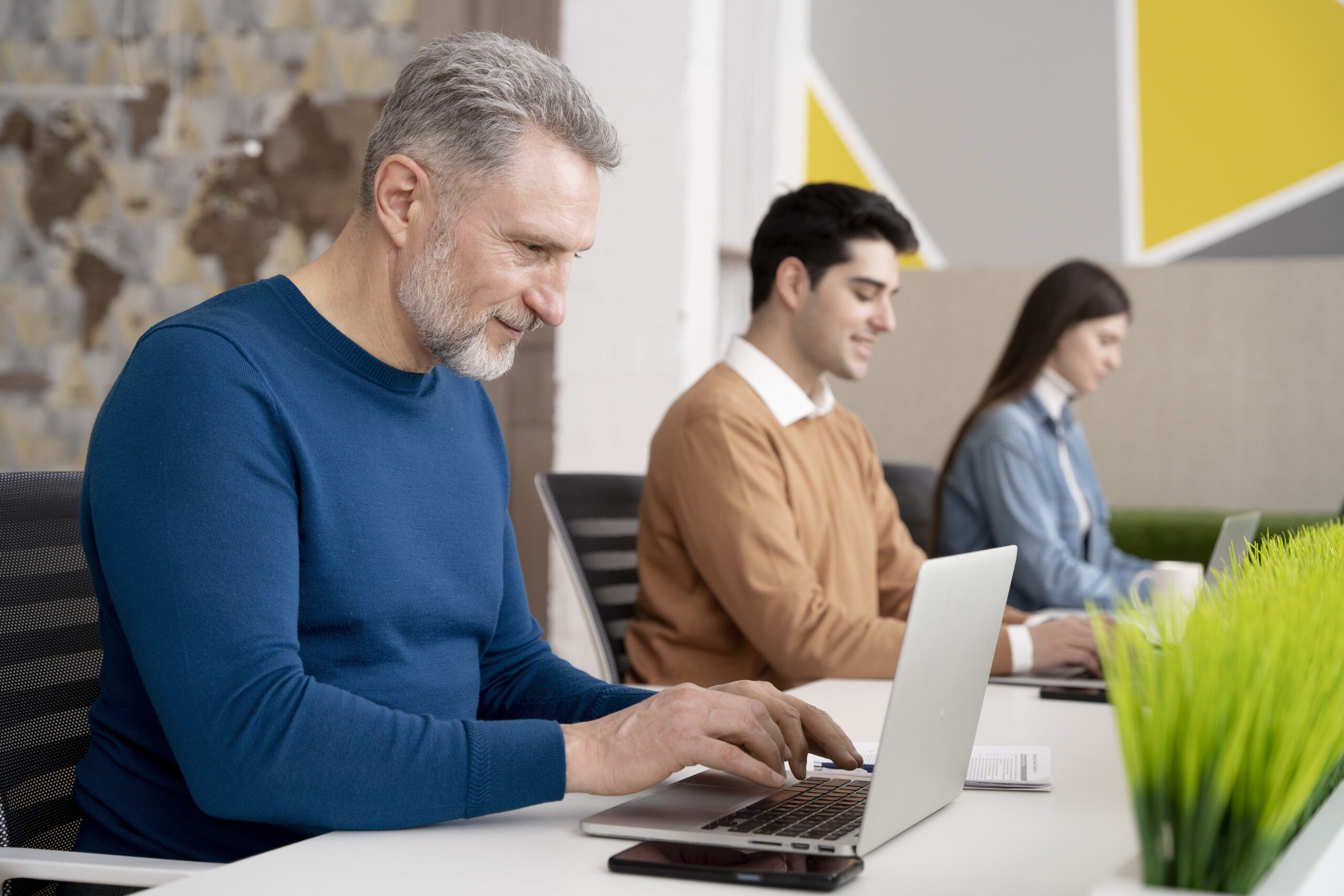 A smiling man with gray hair works on a laptop at a desk, wearing a blue sweater. His two colleagues sit beside him, focused on their laptops discussing outsourced payroll management services. A potted plant decorates the table in this modern office setting.