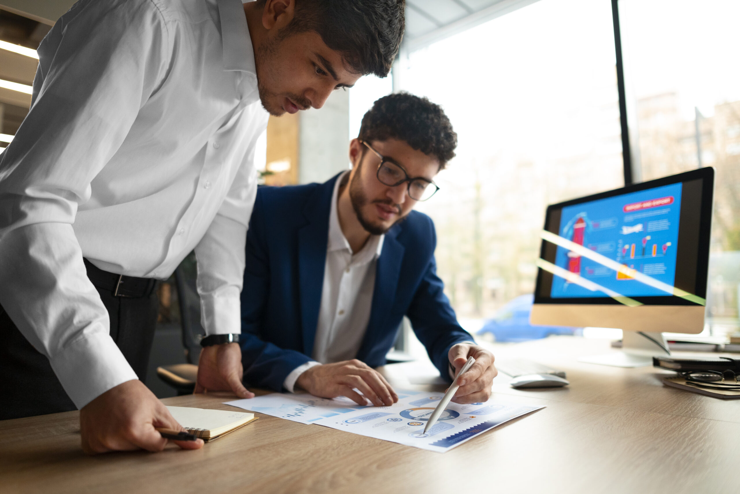 Two men are focused on a document on a desk in an office setting, engaged in tax planning and preparation. One is pointing at charts on the paper, while the other holds a notebook. A computer monitor with a chart is in the background, and large windows let in natural light.