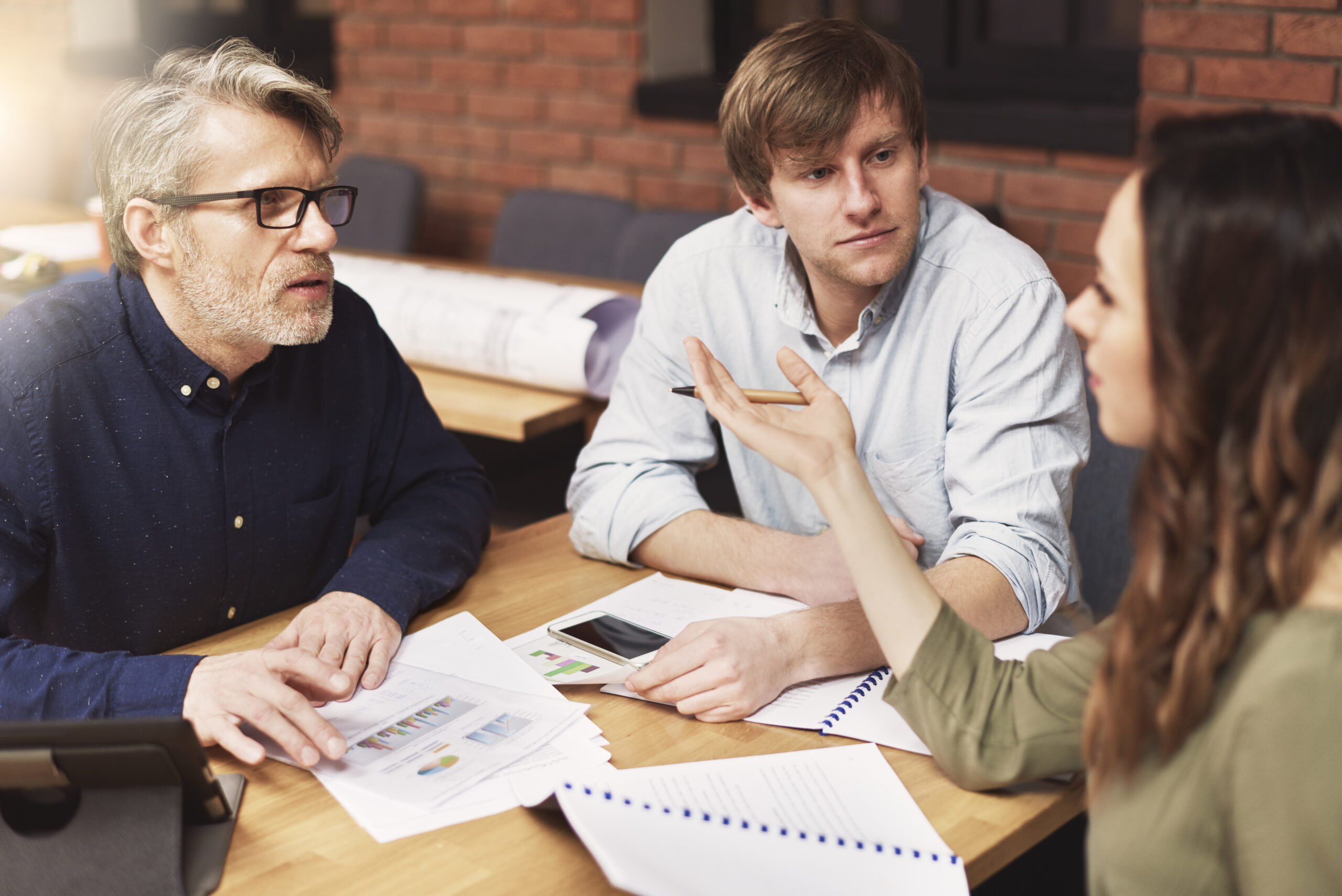 Three people sit at a table engaged in a discussion about an outsourced tax resolution service. Papers, a smartphone, and a tablet are spread out before them. The woman gestures while talking to the two men, who listen attentively with a brick wall in the background.